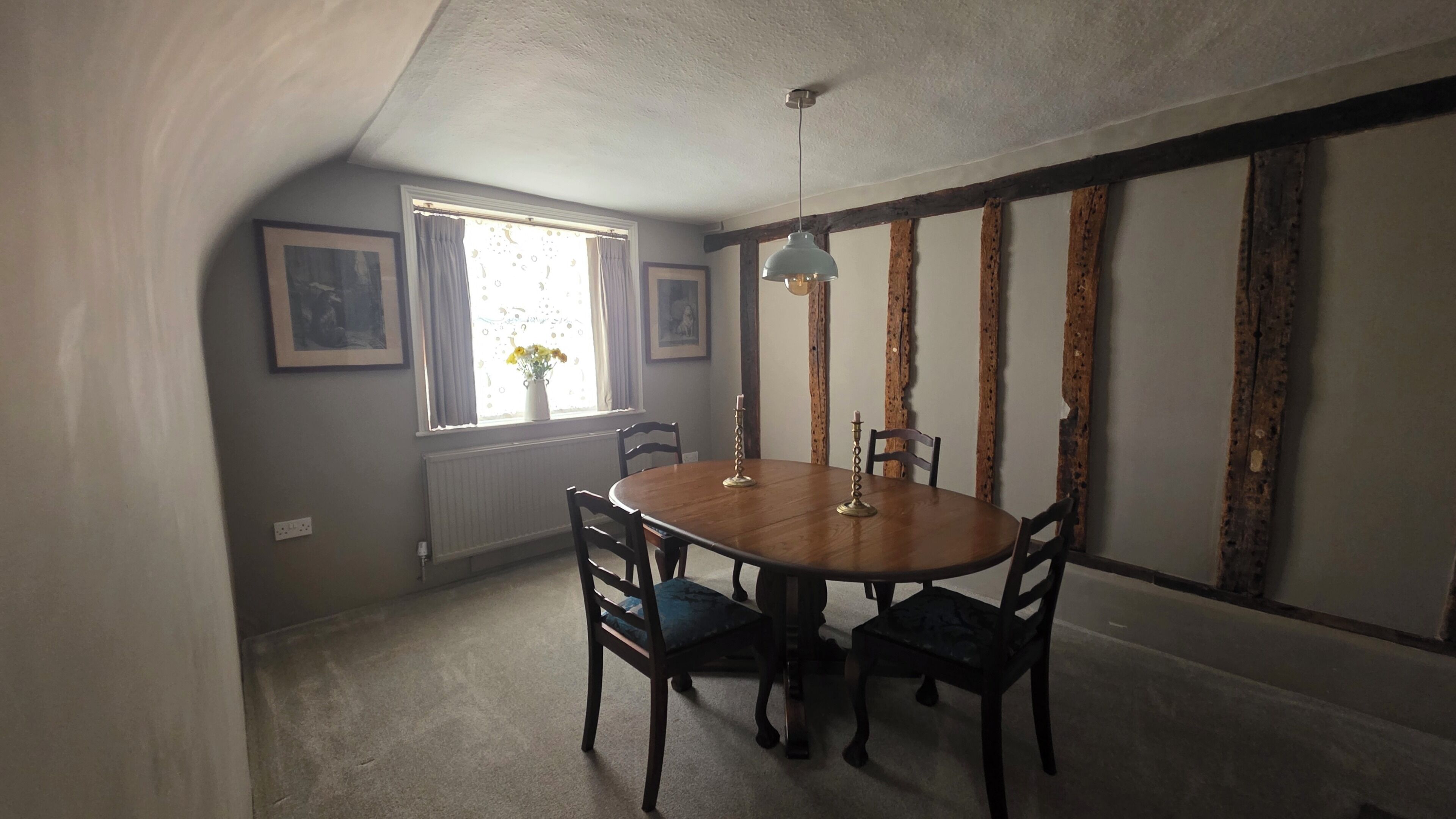 Una sala da pranzo con un tavolo rotondo, quattro sedie, una lampada a sospensione, una finestra con tende e un termosifone.