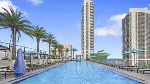 Una piscina con sillones de descanso y sombrillas, rodeada de edificios altos y palmeras.