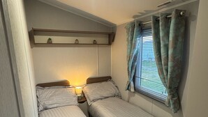 Une petite chambre agréable, dotée de deux lits simples, d’une table de chevet avec une lampe, d’une étagère ornée d’objets décoratifs et d’une fenêtre avec des rideaux.