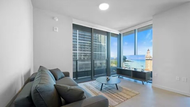 Un salón moderno con un ventanal grande, un sofá, una mesa de centro y vistas a un paisaje urbano.
