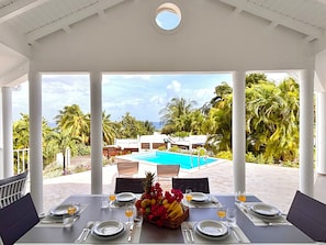 A covered outdoor dining area with a pool view, tables set with plates, glasses, and fruit. - Villa "Les Coraux" (Le Moule)