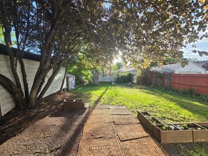 A backyard with a paved walkway, a raised garden bed, and a tree providing shade. - Comfortable 2 Bedroom Upper-Level Home in Sugarhouse (Salt Lake City)