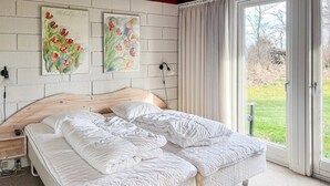 Un dormitorio con cabecera de madera, cama con ropa de cama blanca, dos cuadros en la pared y vistas a través de un ventanal.