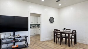 Un salon moderne avec un téléviseur à écran plat, une table à manger avec des chaises, une cuisine en arrière-plan et une horloge au mur.