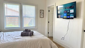 Une chambre à coucher avec un lit, deux fenêtres équipées de stores, un téléviseur fixé au mur qui permet d’accéder à des services de diffusion en continu, et une porte.