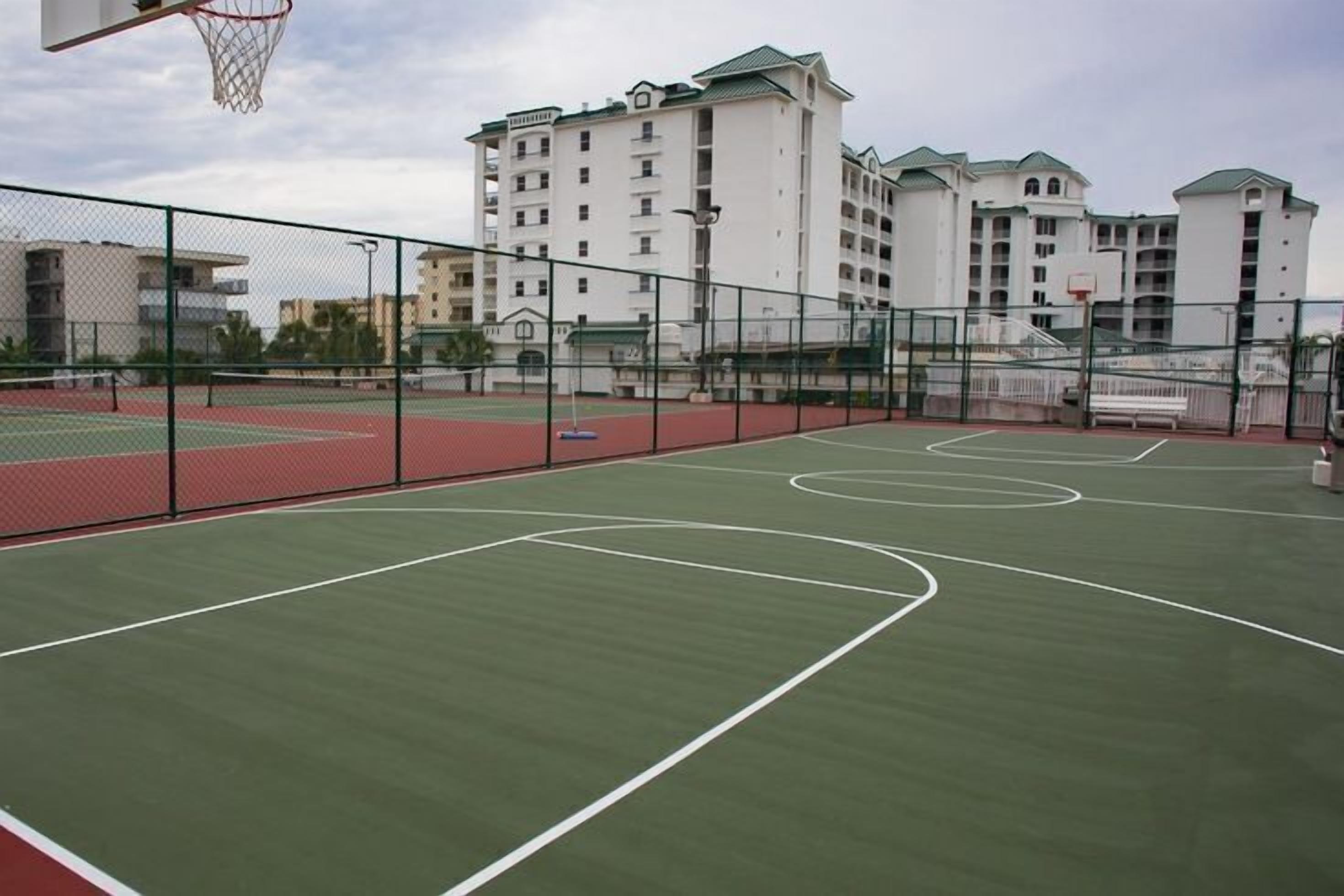Un campo da basket all'aperto con canestro, circondato da una recinzione, con edifici sullo sfondo.