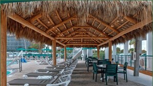 A covered outdoor area with lounge chairs and tables, a pool in the background, and a thatched roof structure. - Penthouse Oceanfront Stay in Cocoa Beach (Cocoa Beach)