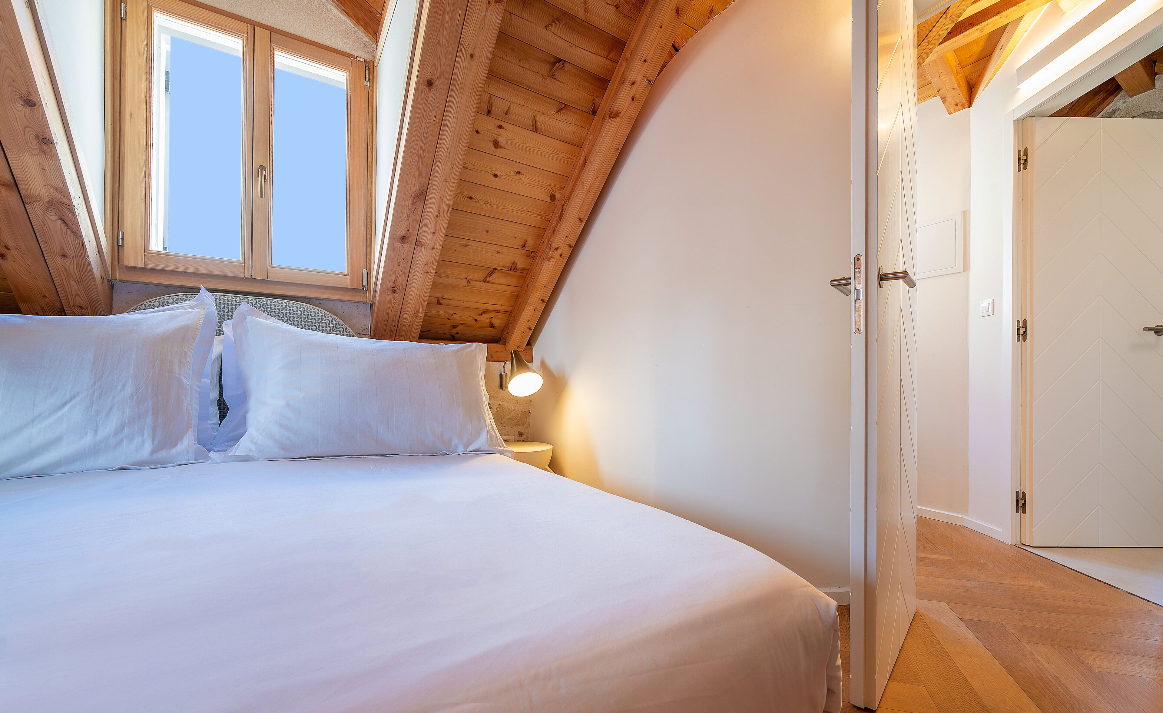 A bedroom with a wooden ceiling, a bed with white bedding, a bedside lamp, and a window.