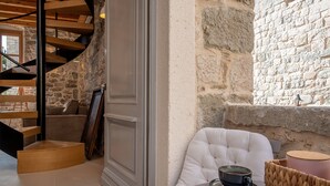 A room with a stone wall, a white cushioned chair, a table with a cup, and a wooden staircase. - Exclusive Heritage Suite Balturio (Kaštel Stari)