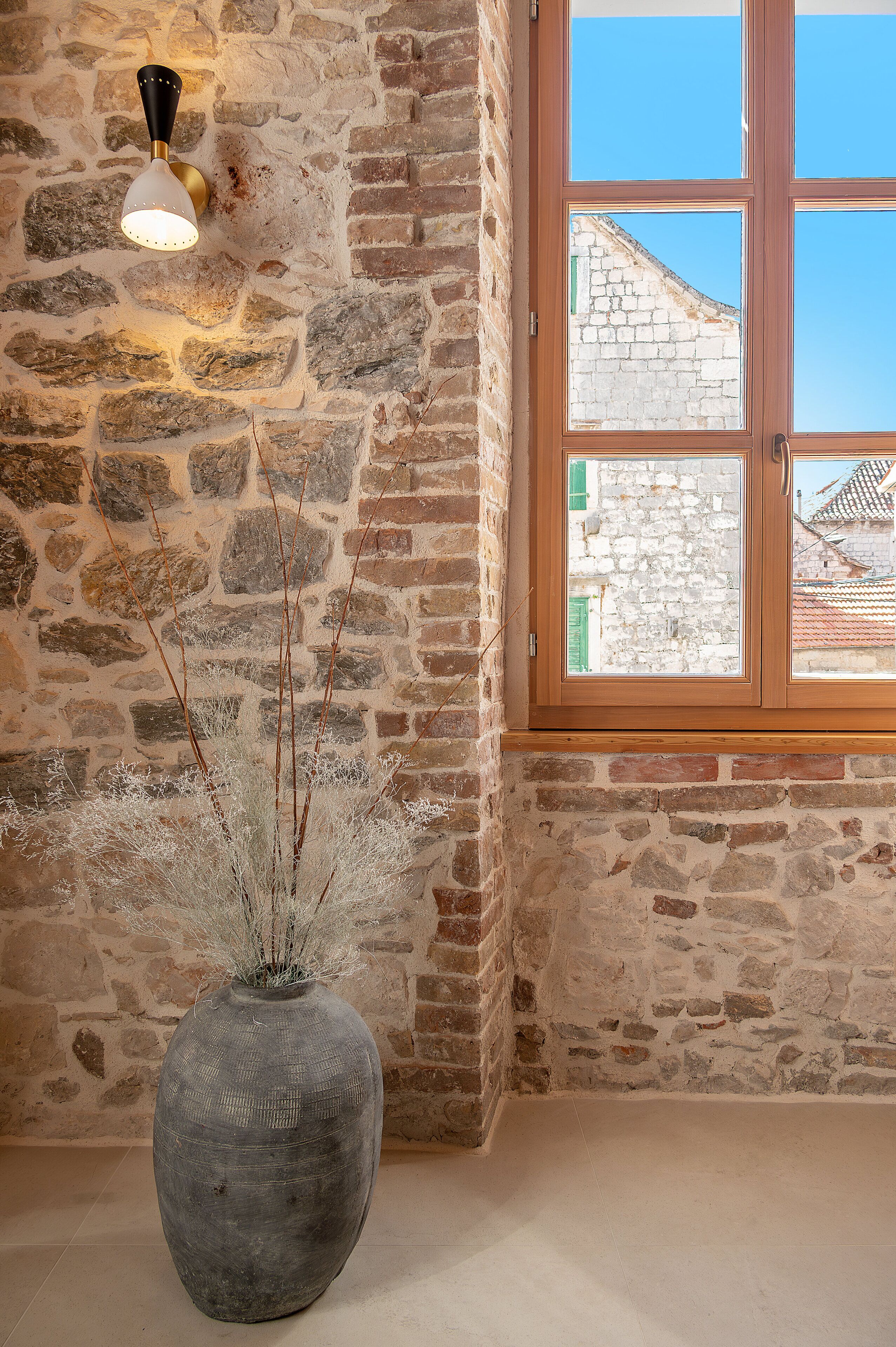 Un muro de piedra con una ventana, un jarrón con ramas y una lámpara fijada a la pared.