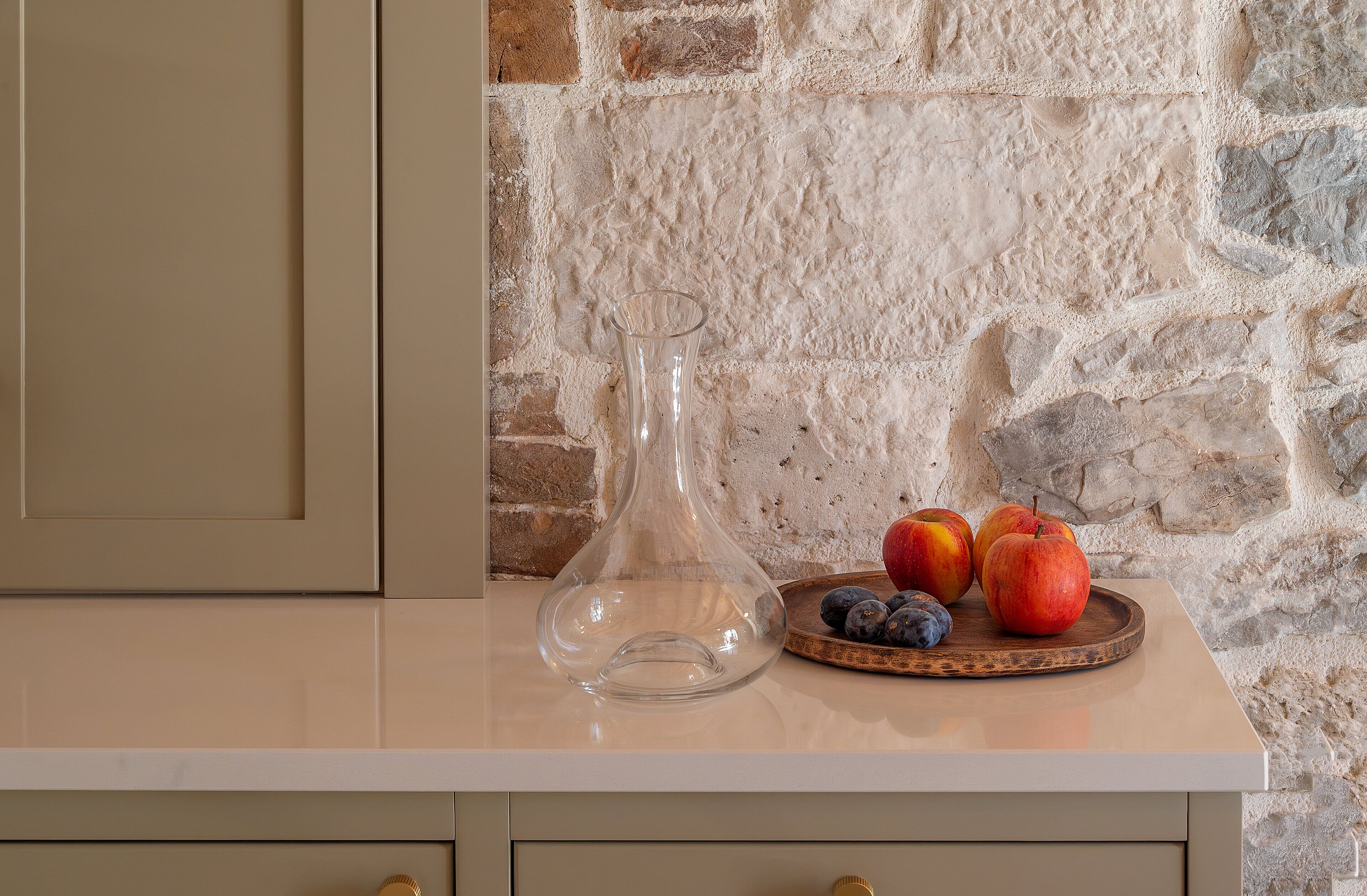 Un comptoir de cuisine avec un vase en verre transparent, un plateau en bois contenant des pommes et des myrtilles, et un mur en pierre en arrière-plan.