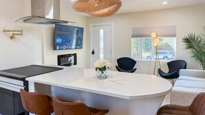 A modern kitchen with a central island, bar stools, a flat-screen TV, and a dining area with chairs.