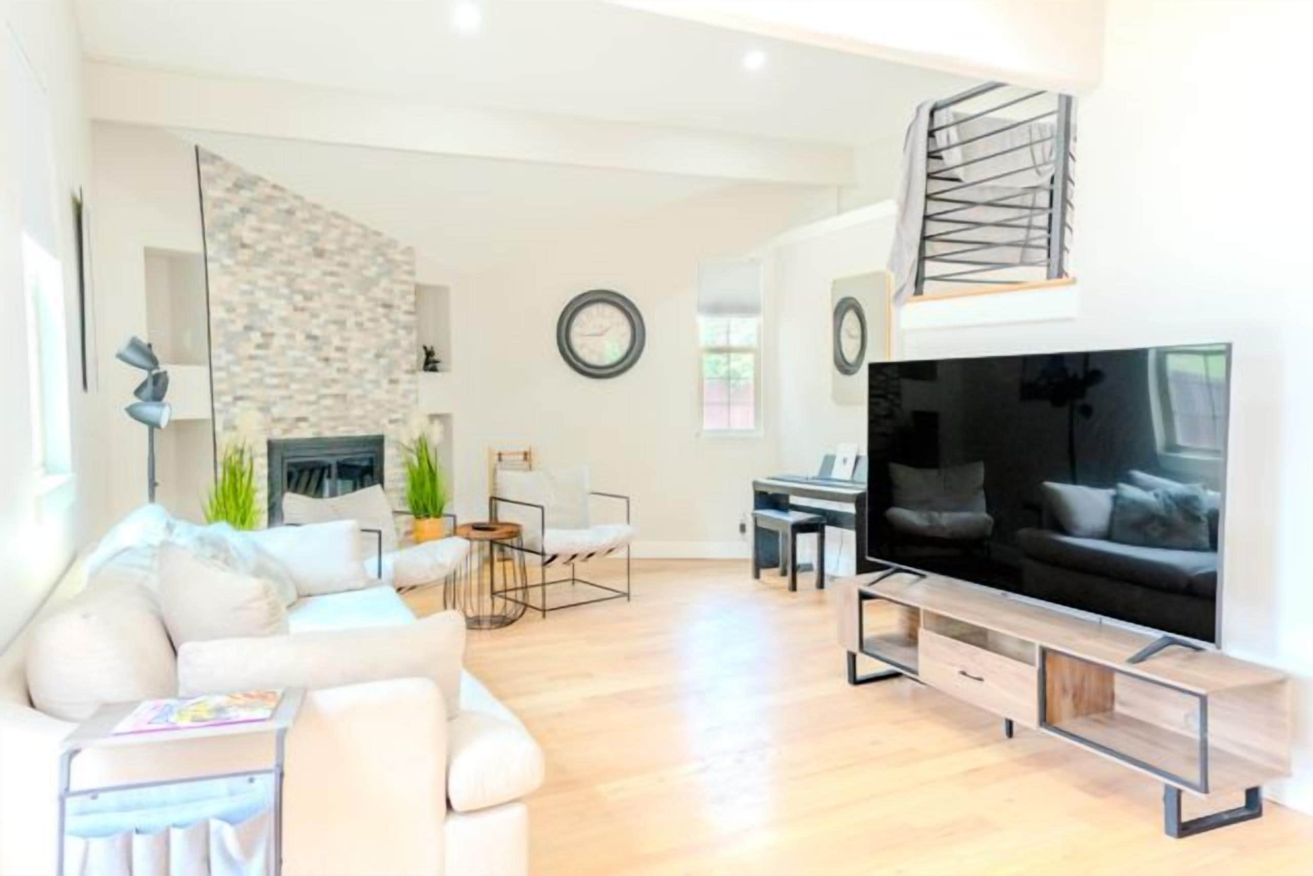 A modern living room with a sofa, coffee table, and a flat-screen TV. There is a fireplace, a dining area, and a staircase.