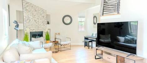 A modern living room with a sofa, coffee table, and a flat-screen TV. There is a fireplace, a dining area, and a staircase.