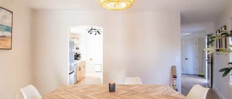 A dining area with a wooden herringbone table, white chairs, a hanging chandelier, and a view into the kitchen.