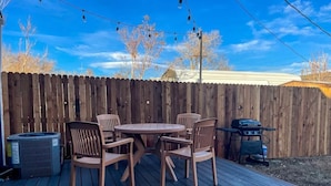Een houten terras met een ronde tafel en stoelen, een barbecue en een afvalbak.