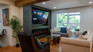 Una sala de estar con televisor de pantalla plana, chimenea, sofá, sillón y ventana con vista a árboles.