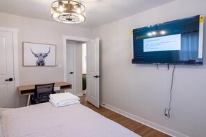 Un dormitorio con una cama, un escritorio, una silla, un televisor montado en la pared y una puerta que da a otra habitación.