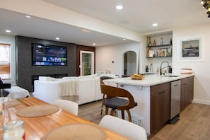 A modern kitchen with a central island, a dining area with a wooden table, and a living area with a sofa and TV.