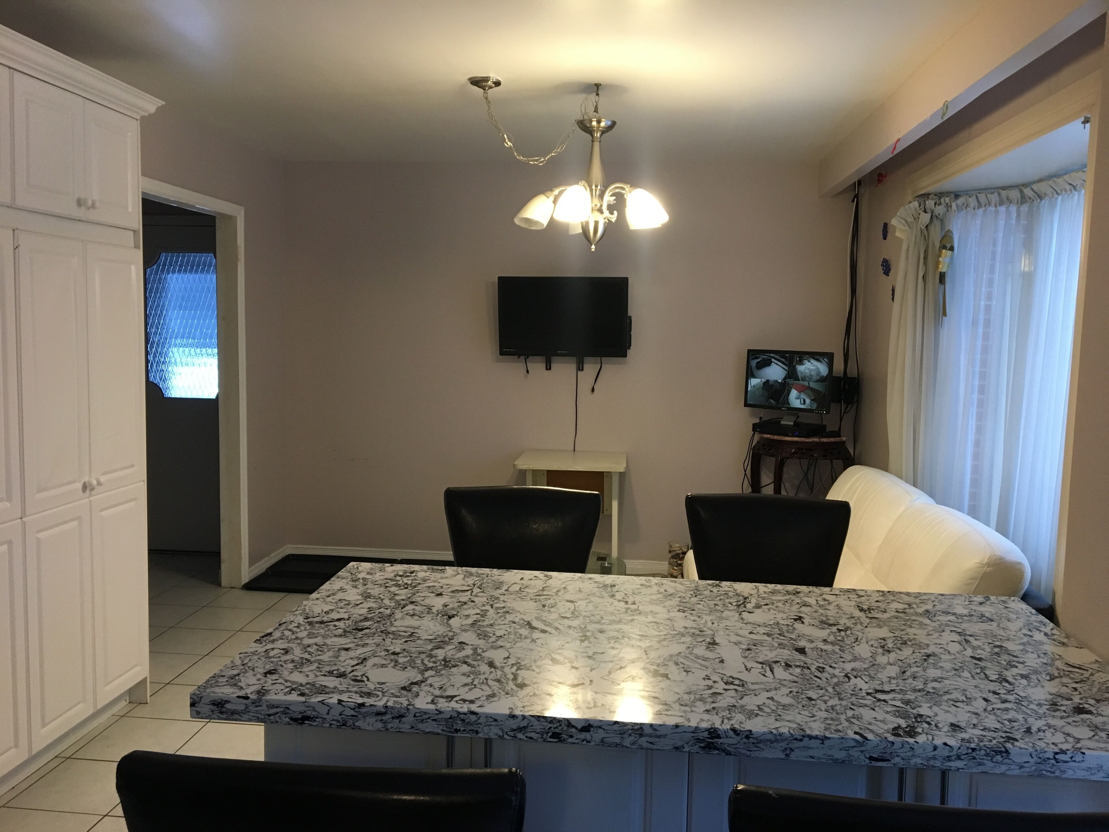 A dining area with a marble table, black chairs, a ceiling light fixture, a flat-screen TV, and a window with curtains.
