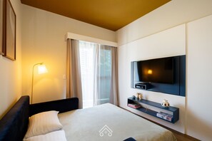 A hotel room with a bed, a TV mounted on the wall, a bedside table with a lamp, and a window with curtains. - Cozy Studio Itaim Bibi (São Paulo)