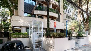 A modern building with a glass elevator, a car parked in front, and a staircase leading to the entrance. - Studio with Parking Space in the Heart of Itaim (São Paulo)