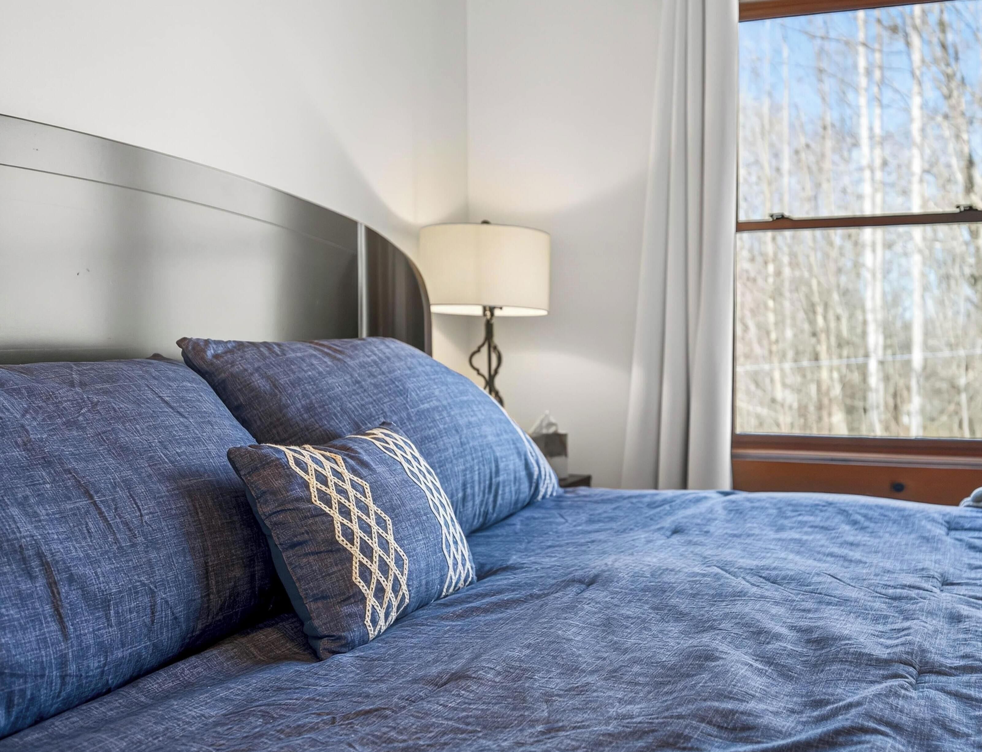 Una cama con ropa de cama azul y almohadas estampadas, una lámpara de noche y una ventana con vistas a árboles.