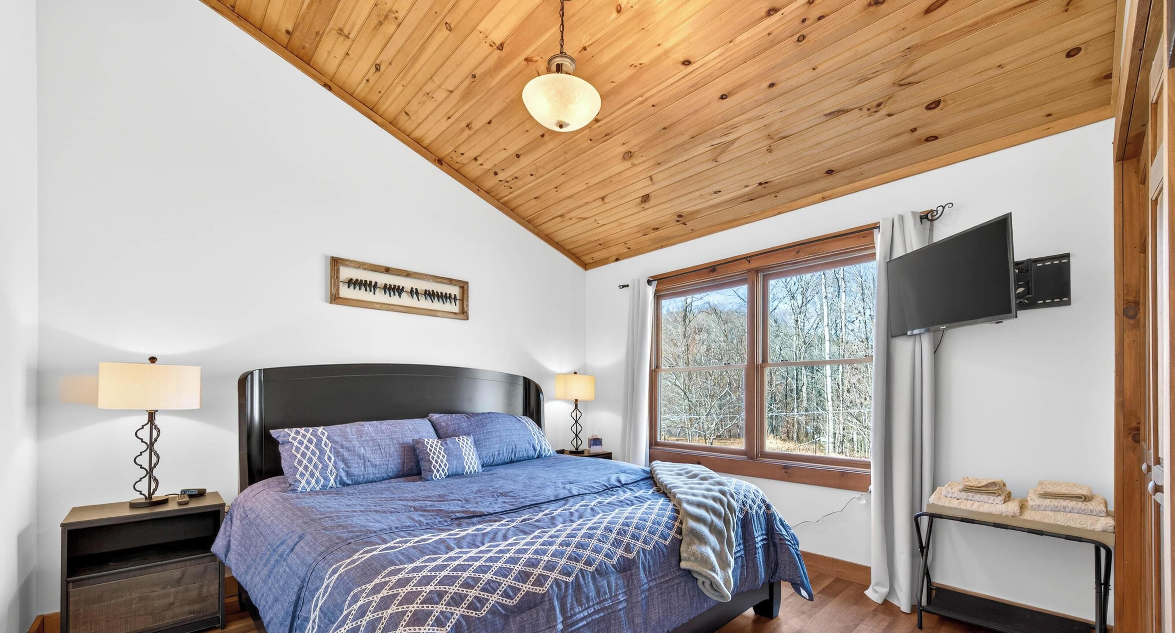 A bedroom with a wooden ceiling, a bed with a blue bedspread, a TV on the wall, and a window with a view of trees.