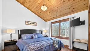 Une chambre avec un plafond en bois, un lit recouvert d’une couverture bleue, un téléviseur fixé au mur et une fenêtre donnant sur des arbres.