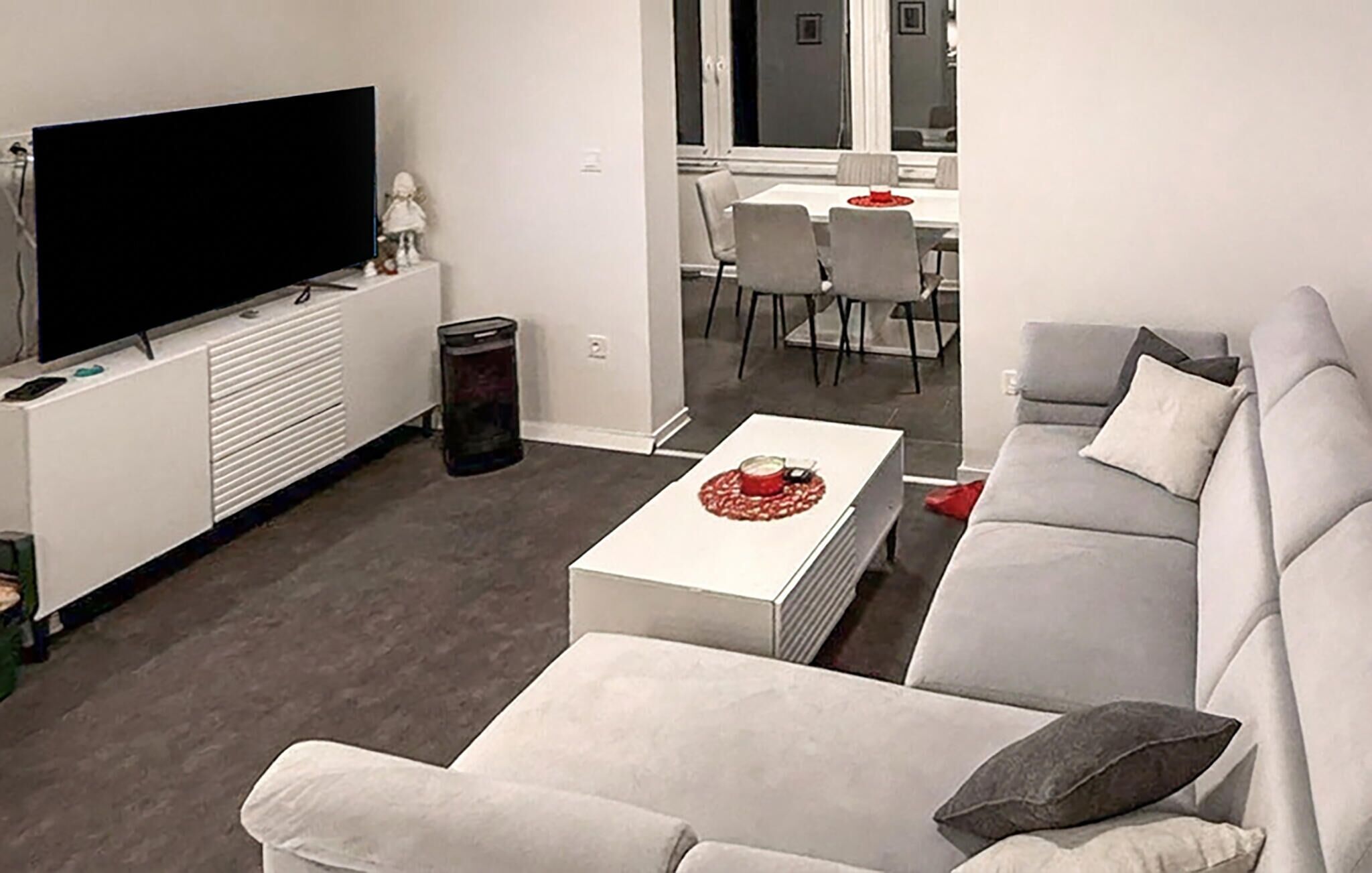 Een moderne woonkamer met een flatscreen televisie, een witte bank, een salontafel met een rode decoratie en een zichtbare eethoek door een open deur.