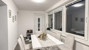 Ein Essbereich mit einem weißen Tisch und Stühlen, einem Fenster mit Jalousien und einem Schuhregal an der Wand.