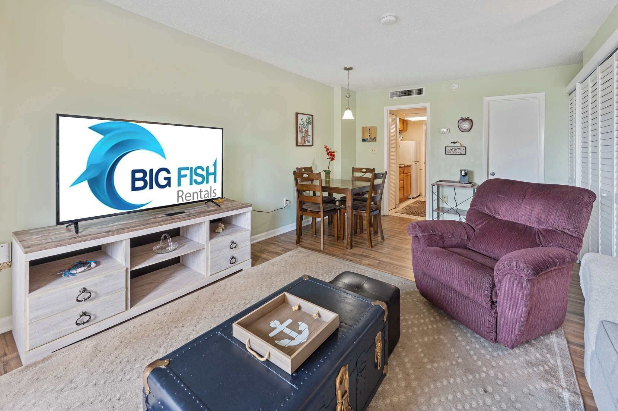 A living room with a television displaying 'BIG FISH Rentals,' a dining area with wooden furniture, and a purple armchair.