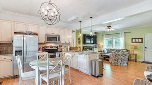 Una cocina moderna con una isla central, comedor con sillas blancas y sala de estar con sofá y televisor.