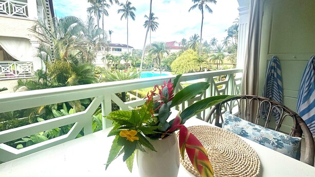 Ein Balkon mit Blick auf einen Pool und Palmen, ein Topf mit rotlaubigen Pflanzen und eine gewebte Tischmatte.