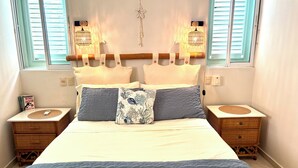 Ein Bett mit einer weiß-blauen Steude, zwei Nachttische mit gewebten Körben, eine Wanddekoration aus Makramee und ein Fenster mit Rollos.