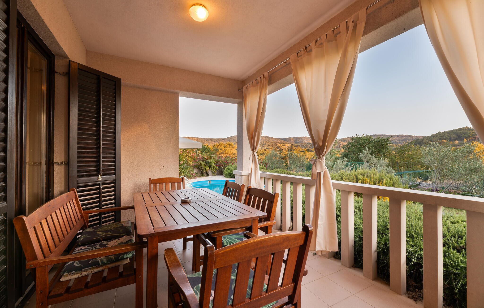 Un balcon avec une table et des chaises, une piscine en arrière-plan et une vue sur un paysage vallonné.