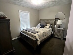 Une chambre à coucher comprenant un lit, une table de chevet, une commode et une fenêtre avec des stores.