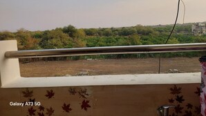 A balcony with a metal railing, a view of a green landscape with trees and buildings in the distance, and a clear sky.