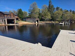 A dock extending into a calm lake with a stone building and a covered area nearby.