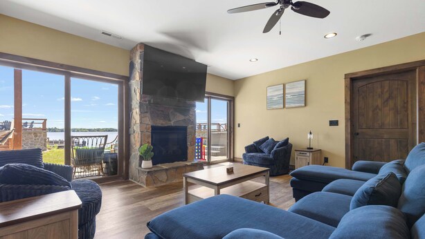 A spacious living room with a stone fireplace, a ceiling fan, and a view of the outdoors. - Lake Front | Pontoon | Private Beach | Hot Tub (New Lisbon)