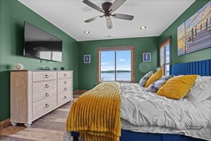 A bedroom with a bed, a dresser, a ceiling fan, a TV, and a window with a view of a body of water.