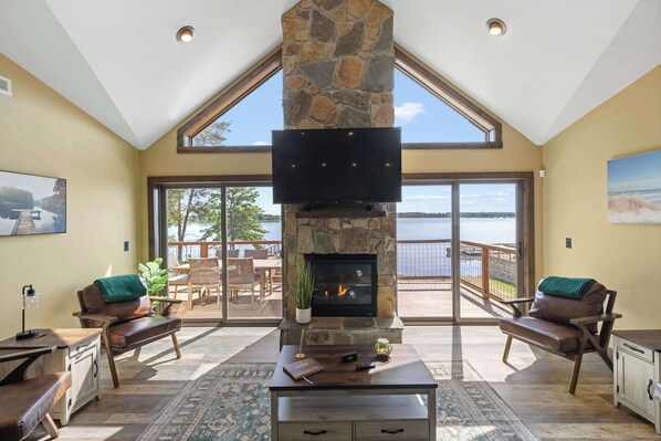 A spacious living room with a stone fireplace, a flat-screen TV, and a view of a lake through large sliding glass doors.