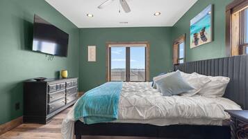 A bedroom with a bed, a dresser, a ceiling fan, a TV, and a window with a view.