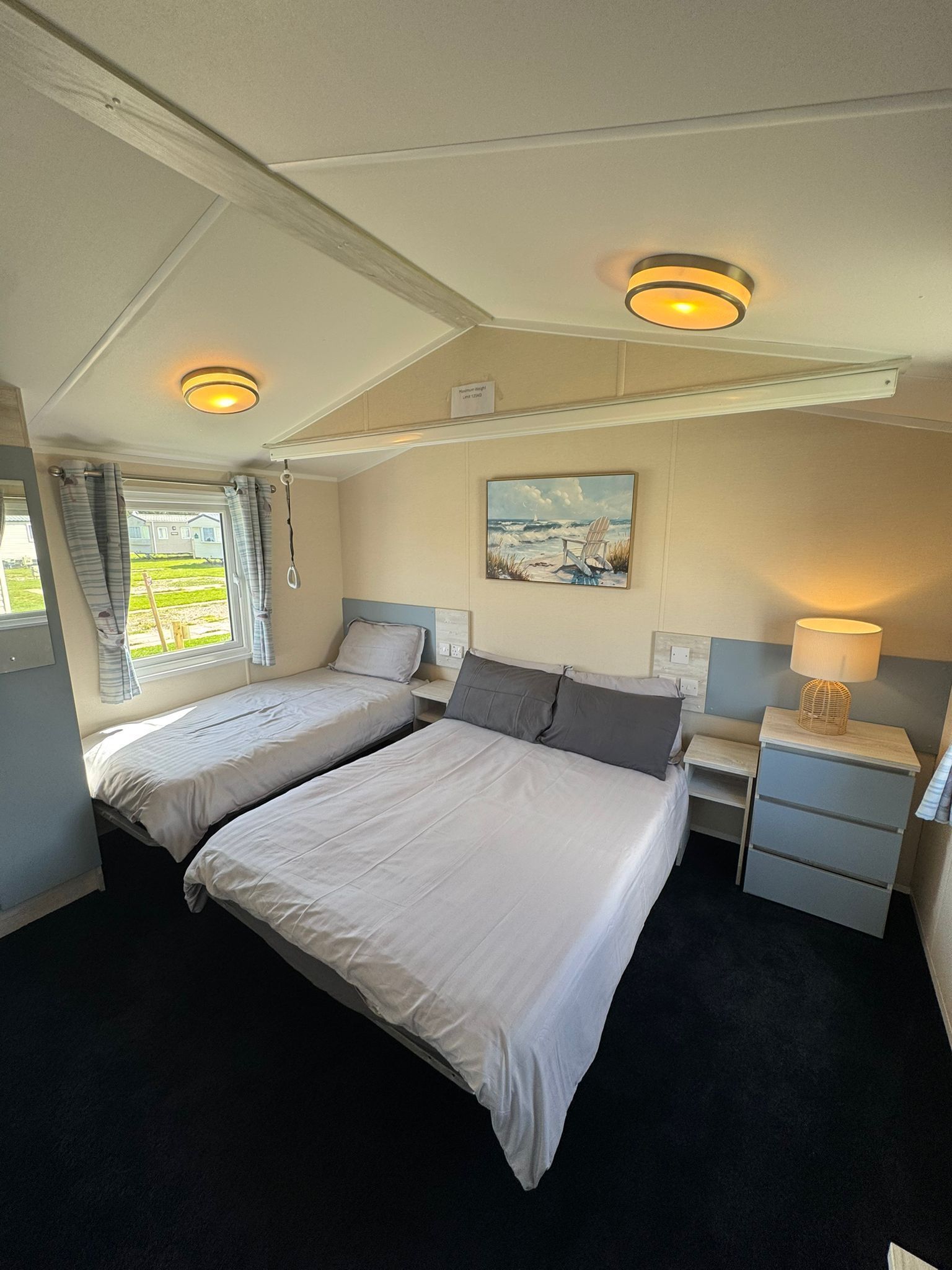 Une chambre avec deux lits, une table de chevet, une lampe, un tableau et une fenêtre avec des rideaux.