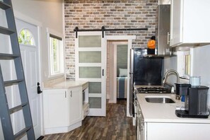 Een keuken met witte kasten, een bakstenen muur, een schuifdeur met glas en een roestvrijstalen koelkast, plus een koffiezetapparaat.