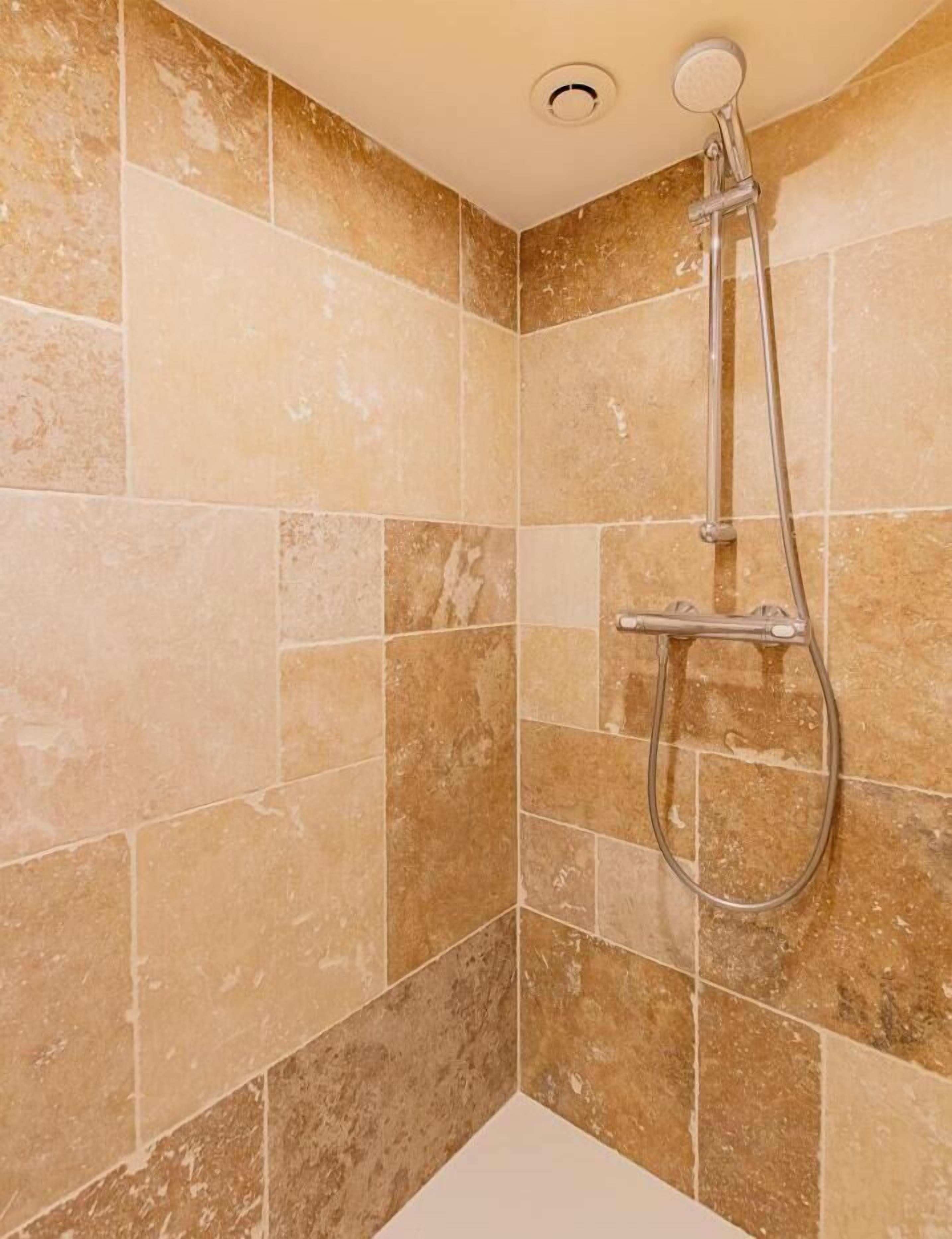 Ducha con cabezal de ducha de mano y grifo empotrado en la pared, ubicada en un rincón con azulejos color beige.