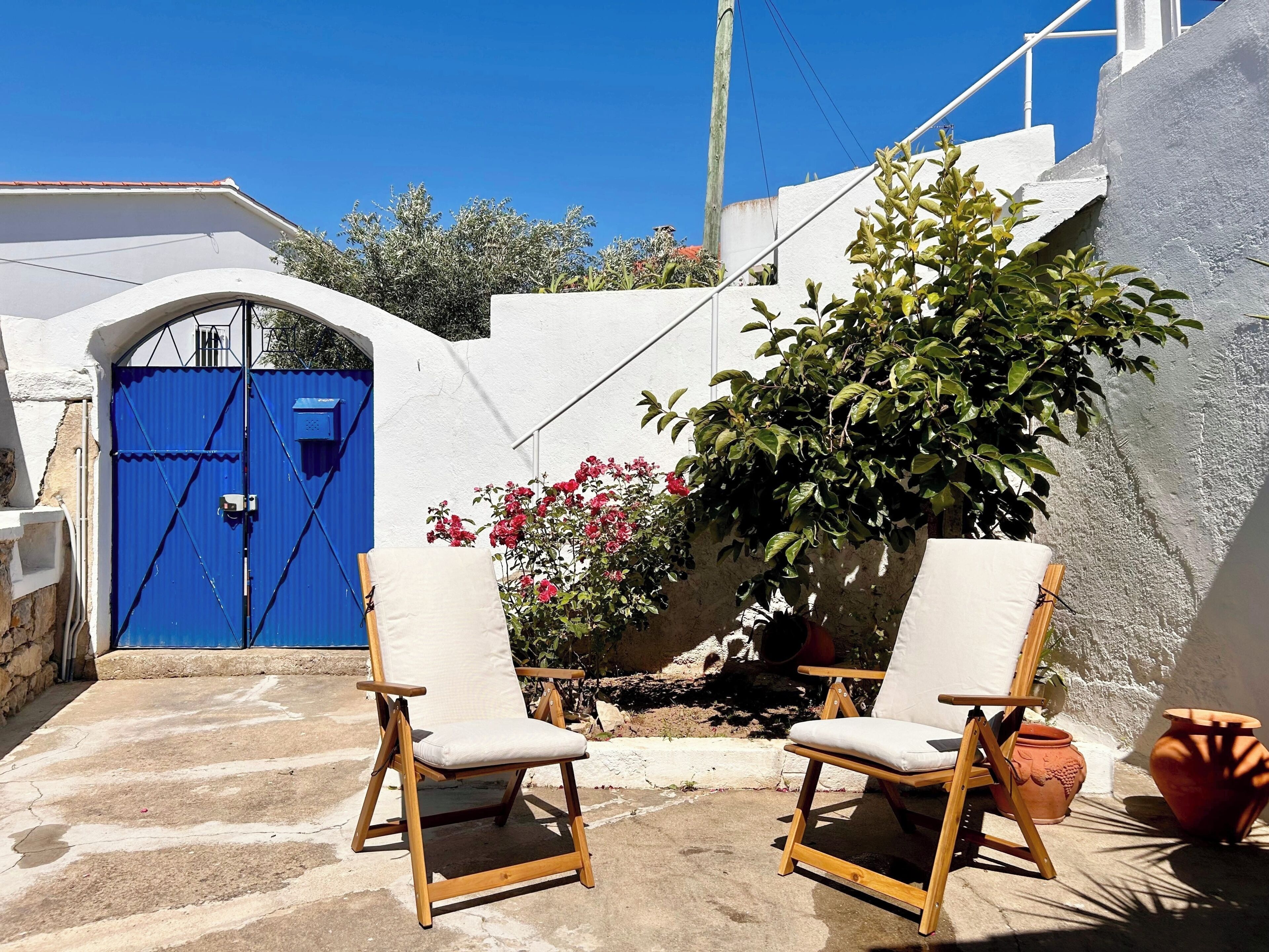 Une cour avec une porte bleue, deux chaises en bois, une plante en pot et des pots en terre cuite.