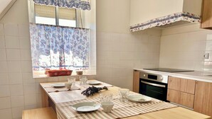 A kitchen with a dining table set for four, a window with floral curtains, and a view of the outside. - Servigueira by Grota Lodging (Leiria)