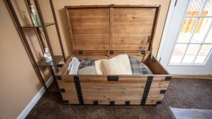 Une boîte en bois dotée d’un couvercle à charnières, posée sur un sol recouvert d’une moquette, à côté d’une étagère en métal où se trouvent des bouteilles.