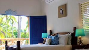 Ein Schlafzimmer mit Bett, Nachttisch, Lampe, Fenster mit Jalousien, Tür und Blick auf Palmen.